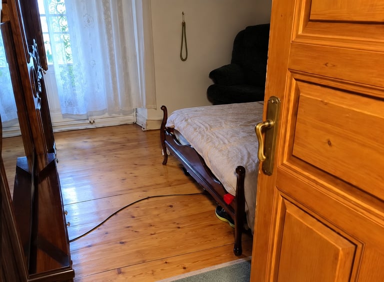 A bedroom with wooden floors and furniture, featuring a bed with white bedding, a wooden dresser, and a window with sheer curtains