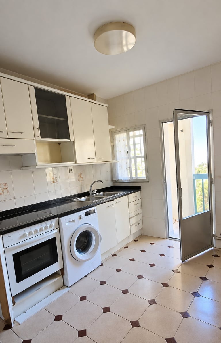 Bright kitchen with cream cabinets, black countertop, white washing machine, built-in oven, and cream and brown tiled floor with glass door to patio